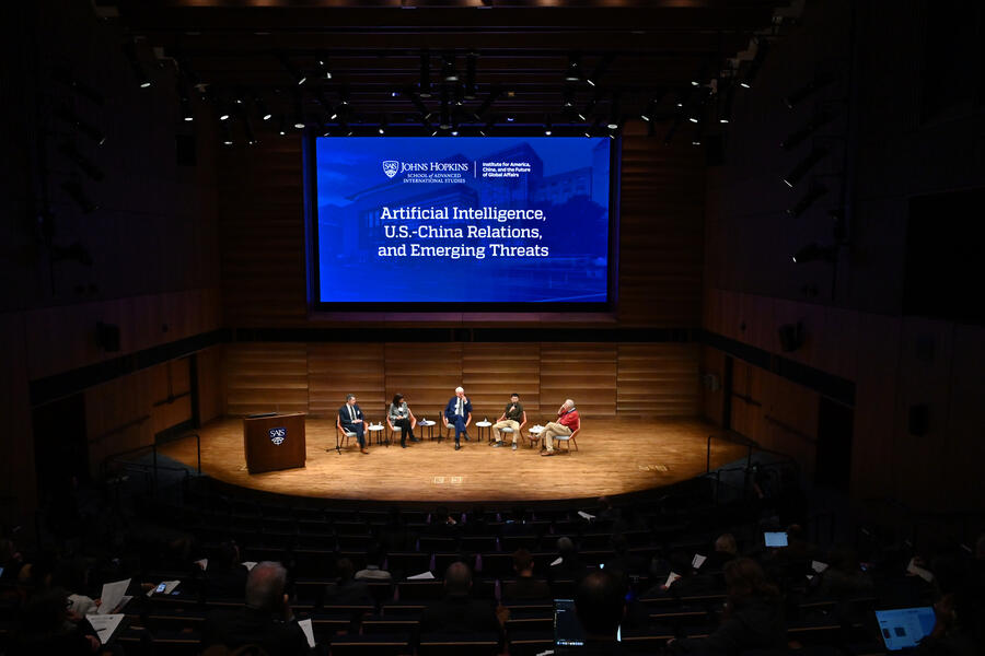 Five speakers sit on a spotlighted stage; above them, a blue screen with the Johns Hopkins logo at the top reads, 