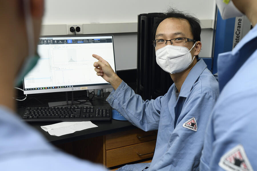 Xiongyi Huang points to a computer screen, looking back to talk to two other people. He is wearing a disposable face mask and a blue lab coat.