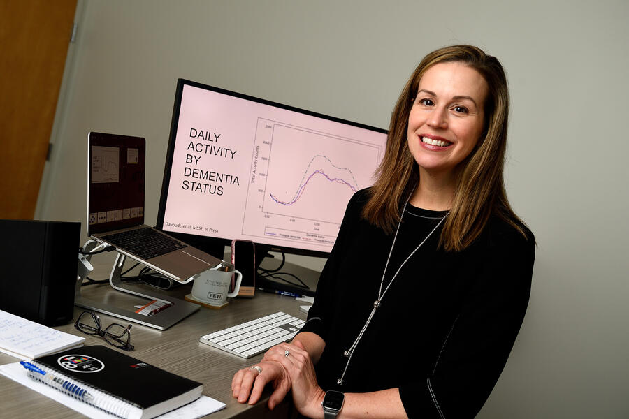 A woman in a black dress smiles while posing for a photo; she sits next to a computer screen that charts 