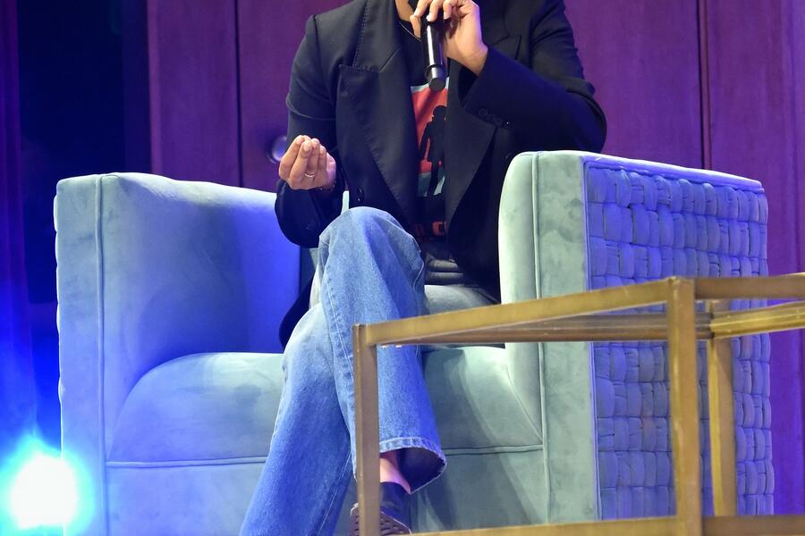 Yara Shihidi sits on a chair onstage while speaking into a microphone. She is dressed in jeans and a blazer.