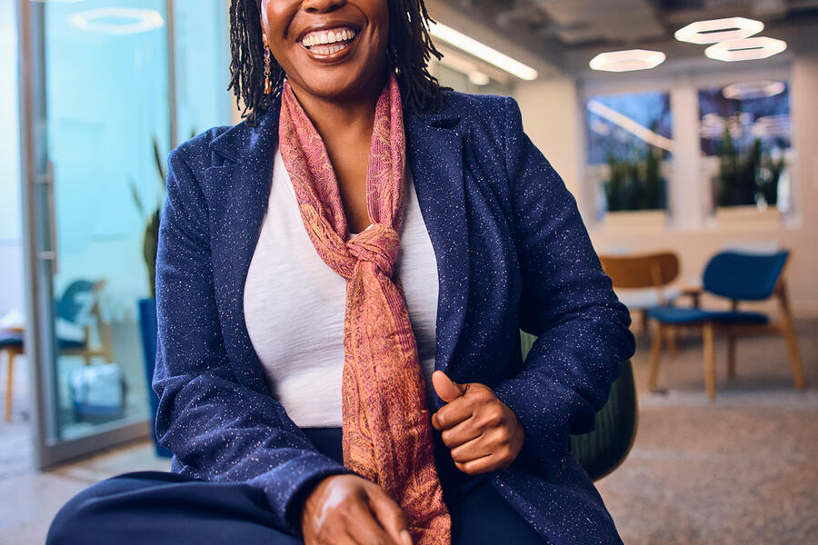 A woman laughs and smiles; she is seated, her legs crossed casually, wearing a white shirt, blue blazer and light red scarf