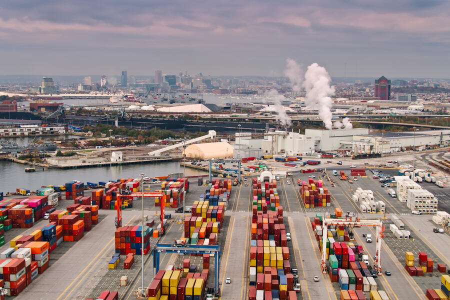 Aerial shot of the Port of Baltimore at sunset, looking across a container terminal and over industrial facilities along the quayside towards the city skyline.