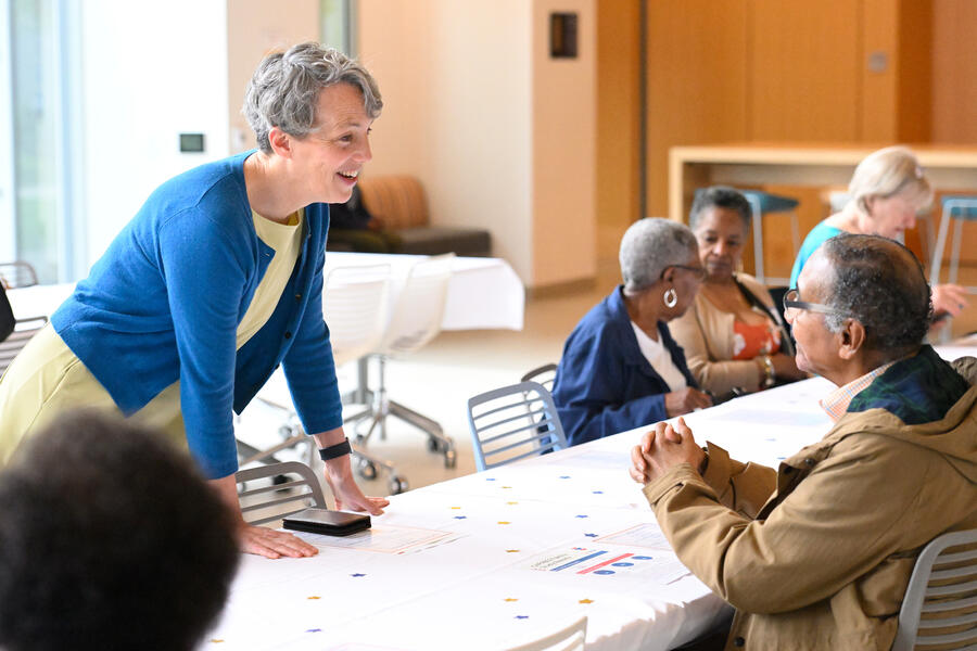 A woman in a yellow dress and blue cardigan speaks with a man who is seated at a table