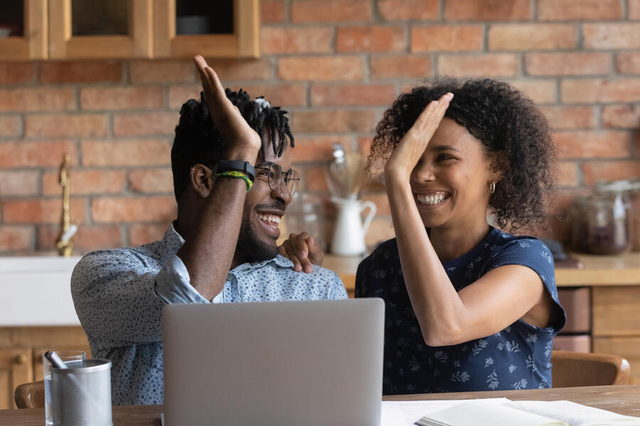 Young couple sitting at a laptop in their kitchen high five each other to show their happiness with what they've acheived