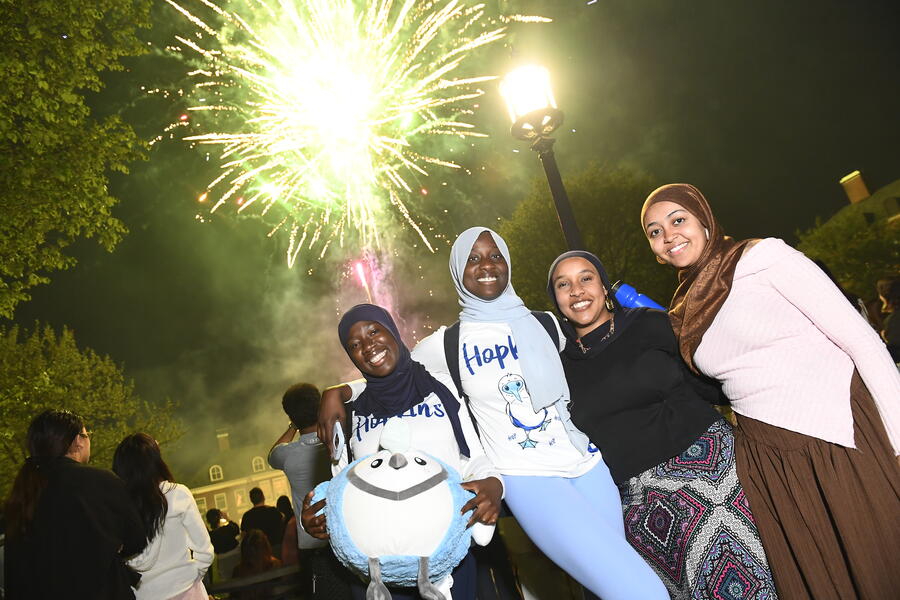 A group of four college students smile for the camera as fireworks go off behind them.