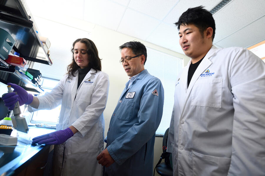 Three people in a lab setting; one is wearing a light blue lab coat, and the other two are wearing white lab coats