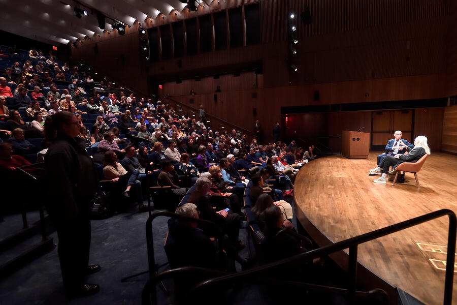 Two people sit on a stage and converse in front of a full auditorium audience