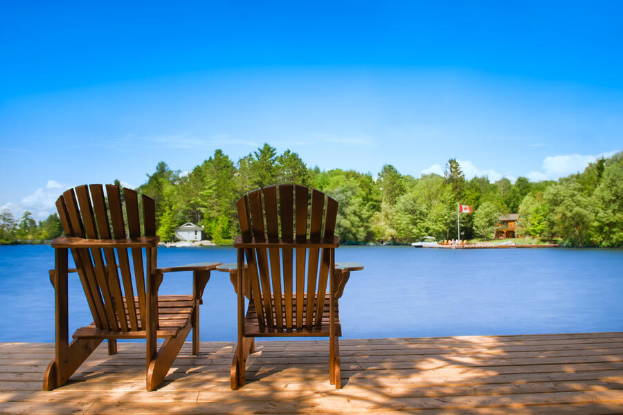 Two empty Adirondacks chairs on a dock facing toward a serene lake and trees