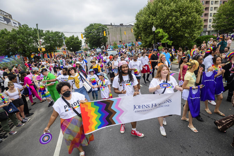 A slightly overhead picture of the 2025 Baltimore Pride Parade. In the front, three people march while holding a sign that says "Johns Hopkins University and Medicine."