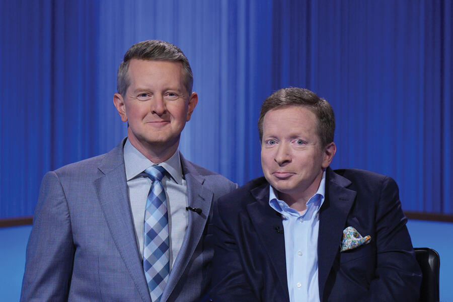 Ken Jennings (left) and alum Harvey Silikovitz stand in front of a blue curtain on the Jeopardy! stage