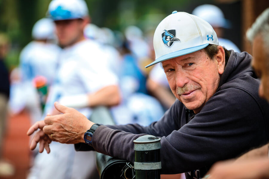 JHU baseball coach Bob Babb leans on a fence while wearing a Blue Jays cap