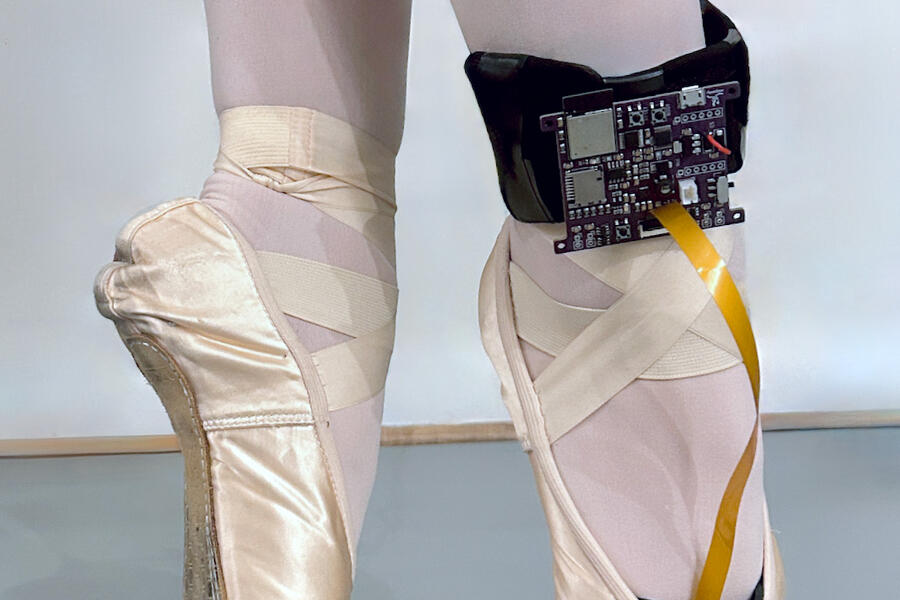 Close-up image of a dancer standing on her toes in ballet pointe shoes; a black control board is wrapped around her right ankle and connected into the toe of the shoe