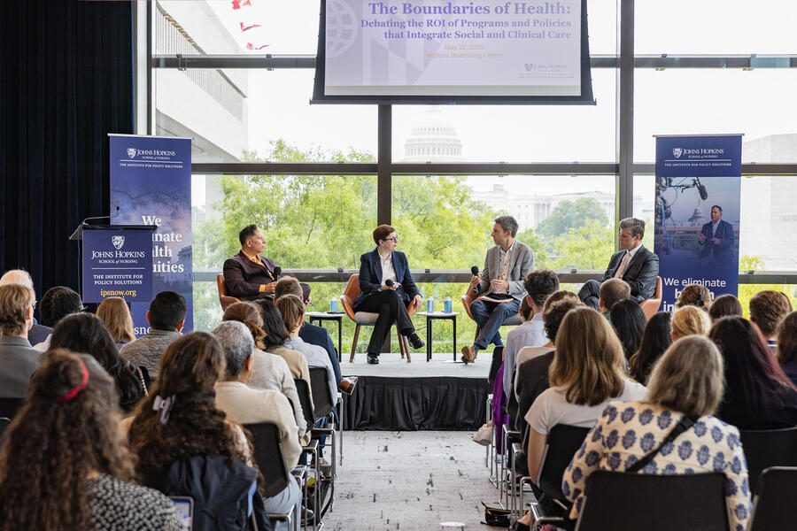 A panel of four people converse on a stage in front of a seated audience; the U.S. Capitol dome is visible through the large windows behind them