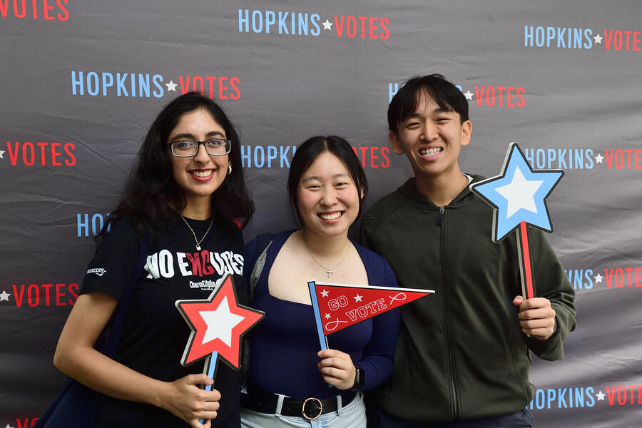 Three students hold up cardboard props--two stars, and a pennant reading "Go Vote"--while smiling.