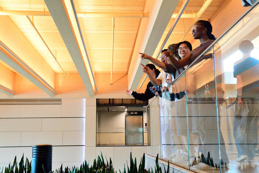 Student stand along an interior balcony in a new building, smiling and pointing out some of the building's features