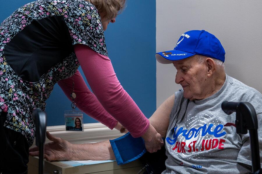 Frank Squibb, one of about 978 people participating in the ARIC study, gets his blood pressure taken by a nurse in colorful scrubs. Frank wears a blue baseball cap and a grey t-shirt.