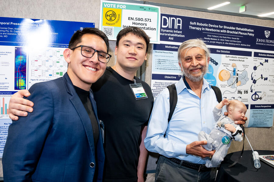From left, Victor Ticllacuri, Yuxuan Bai, and Nitish Thakor pose in front of a poster display for their invention at WSE Design Day 2025. Nitish Thakor is holding a lifelike baby doll, used to demonstrate their invention