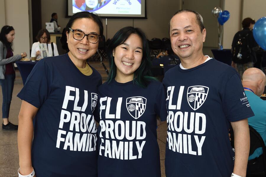 Three people, two older adults and one young adult, smile for the camera while wearing shirts that read "FLI Proud Family."