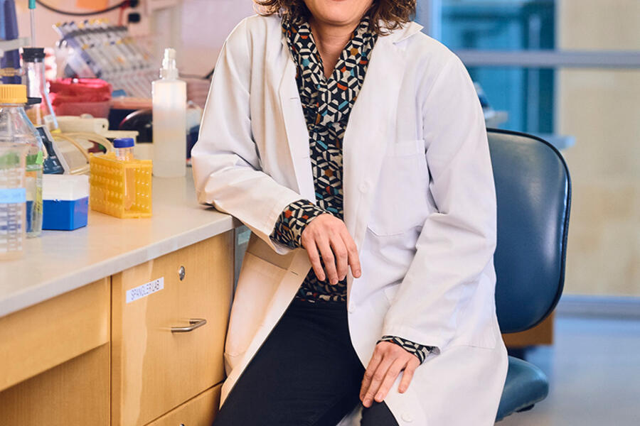 A woman in a white lab coat sits on a chair and poses for a portrait in a lab setting