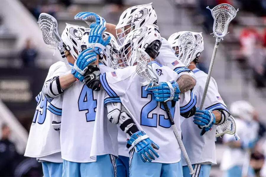 A group of men's lacrosse players in white jerseys and white helmets huddle and celebrate