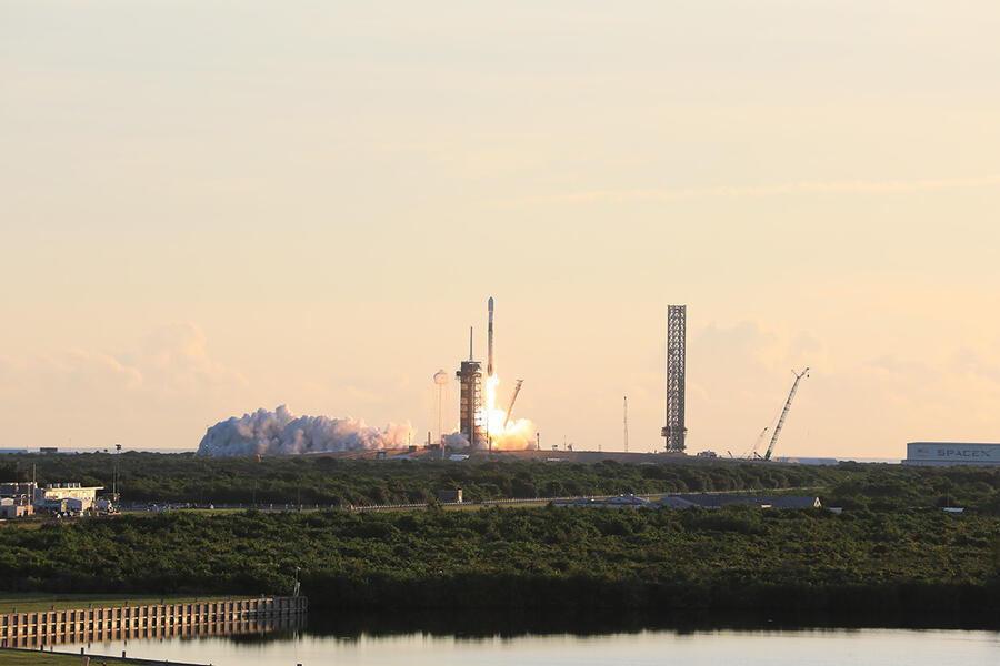 A rocket launches in the distance against a backdrop of a yellow-orange Florida sunrise