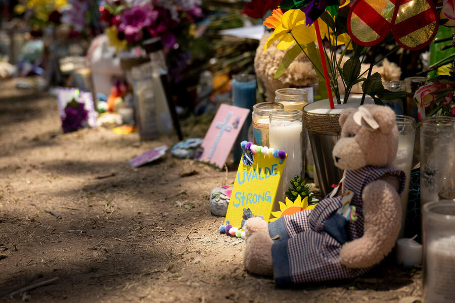 A small teddy bear, flowers, and a sign reading Uvalde Strong sit on the ground in a makeshift memorial