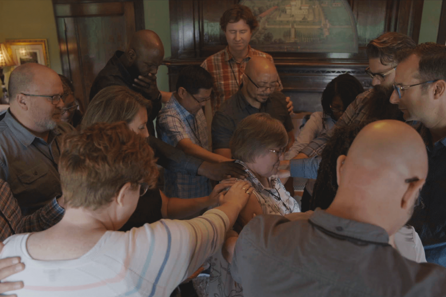 A group of people gather in a circle to pray with their hands on one person's shoulders.