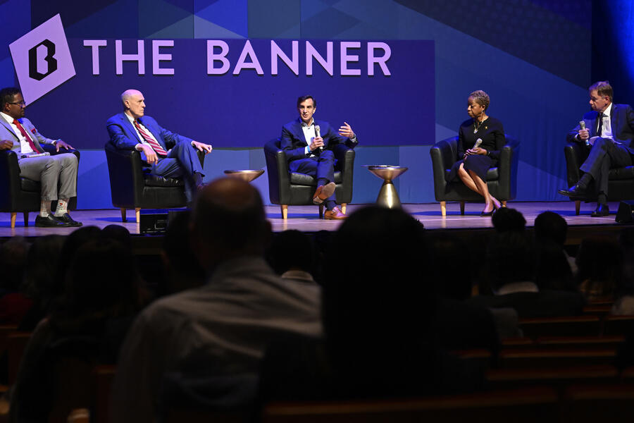 A panel of five individuals sits on stage; A sign reading The Banner is visible in the background