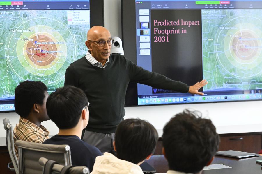 Professor RT Ramesh teaches in front of a class. Behind him is a powerpoint titled "Predicted Impact Footprint in 2031" with an image of a simulated asteroid impact.