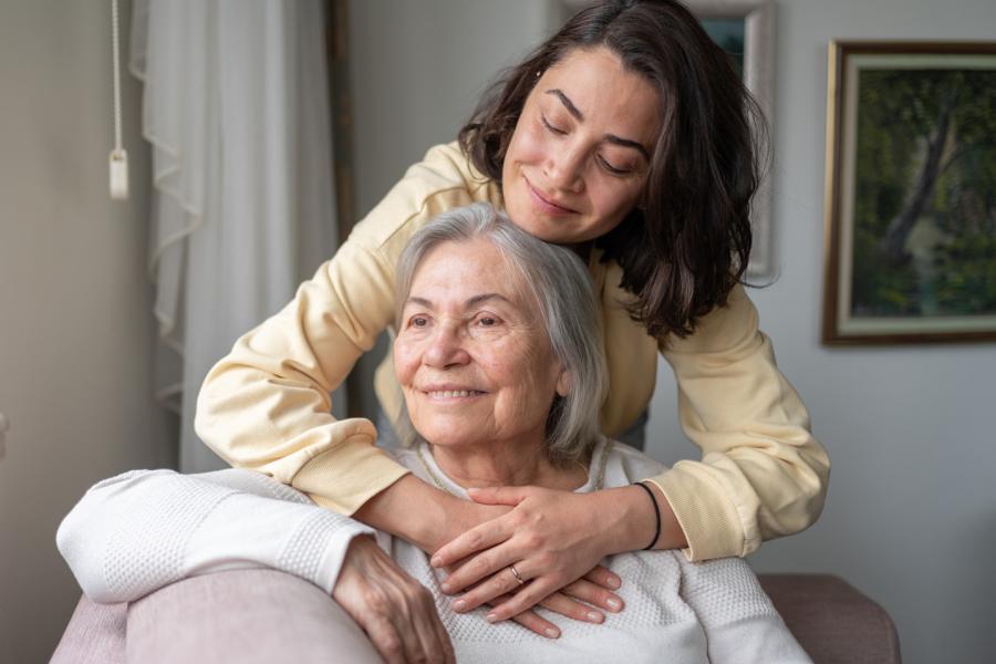 An adult woman hugs her mother, who is seated in front of her.