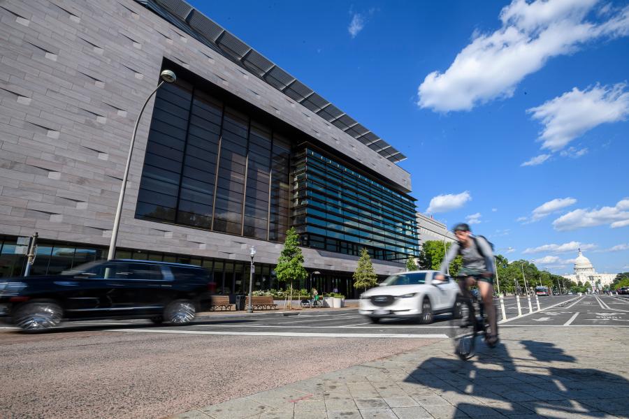 Cars and a cyclist pass a large, gray building