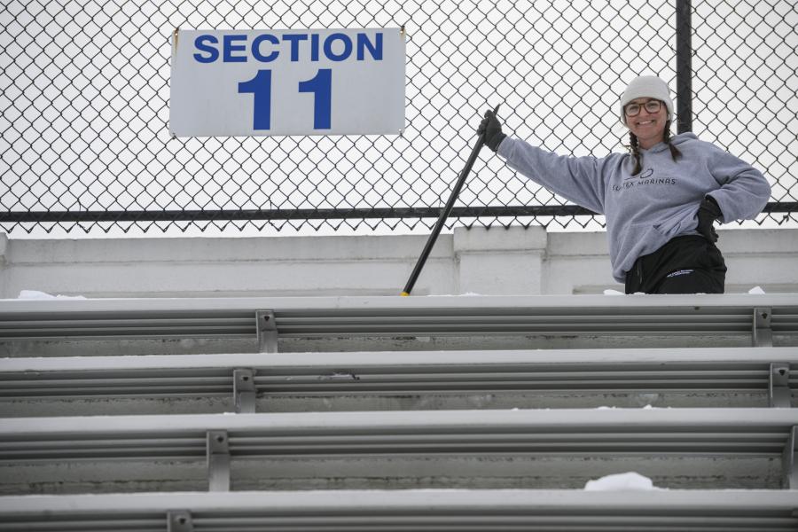 An athletics staff member smiles in the stands at Homewood Field while shoveling snow from the seats