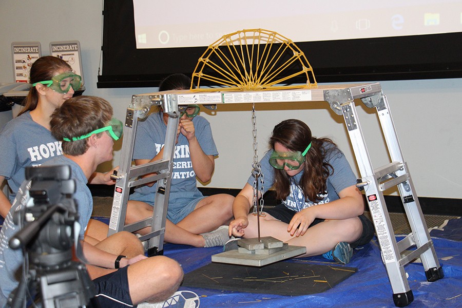 A three-person team tests their bridge with one student gently placing a weight