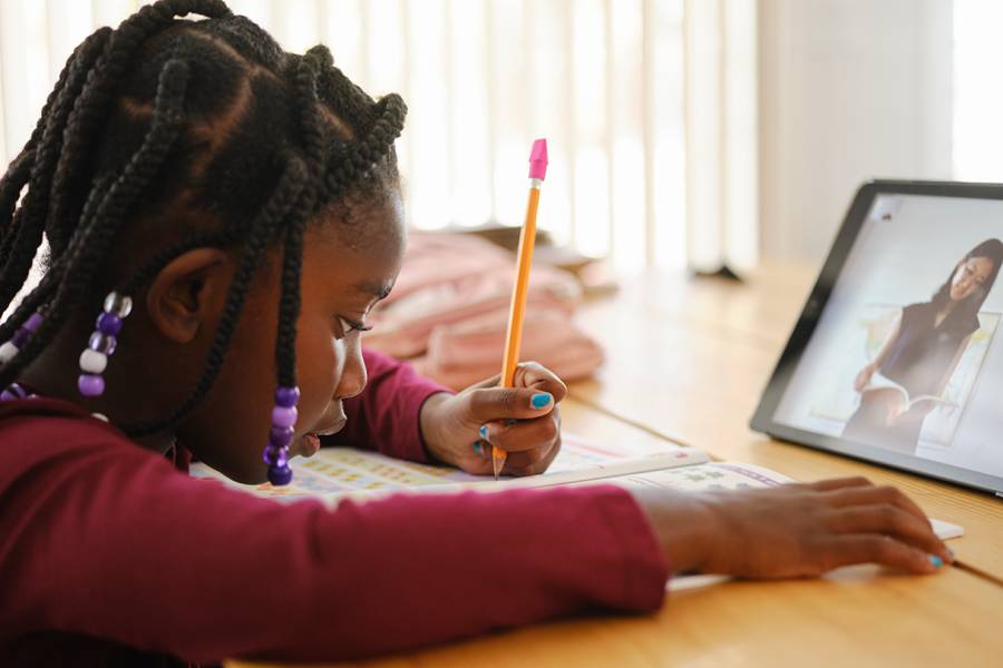 A child does classwork while sitting in front of a tablet displaying a video conference call