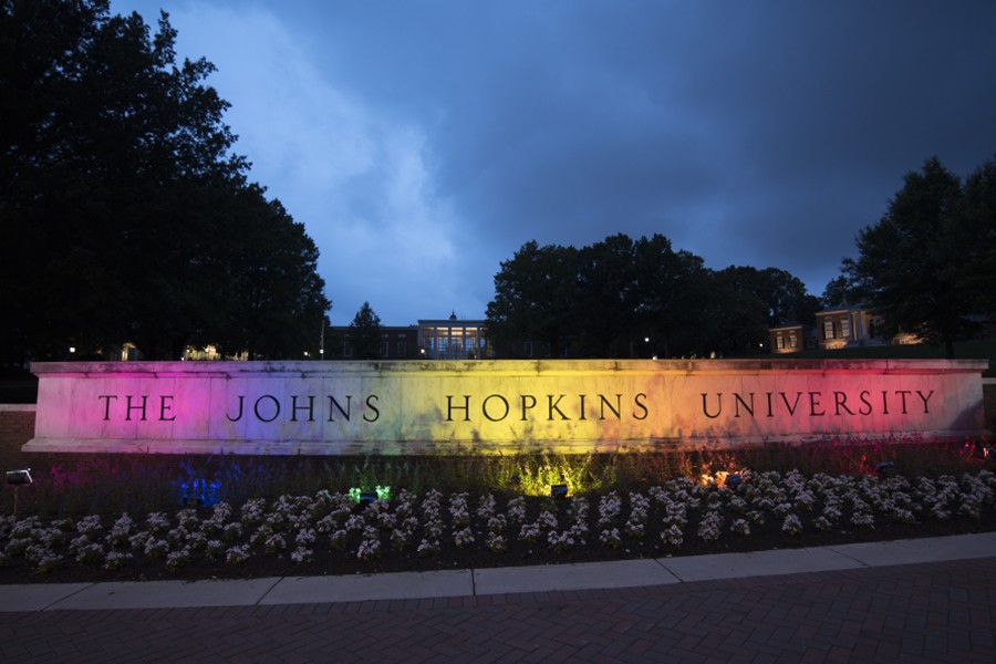 The concrete gateway reading 'The Johns Hopkins University' is lit up with red, orange, yellow, green, blue, and purple lights