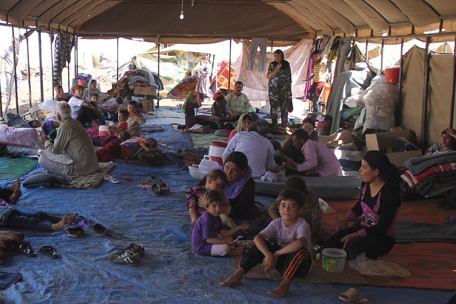 Photo shows people sitting and standing in an open-air tent with makeshift walls from blankets and a dirty tarp floor
