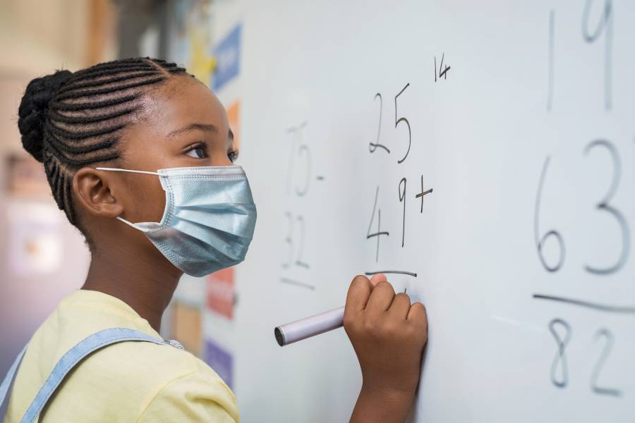A little girl in a mask practices addition on a school whiteboard
