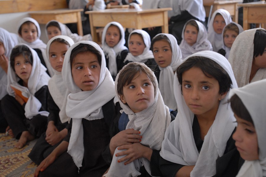 A group of children dressed in black dresses and draped in white head scarves sit in a classroom