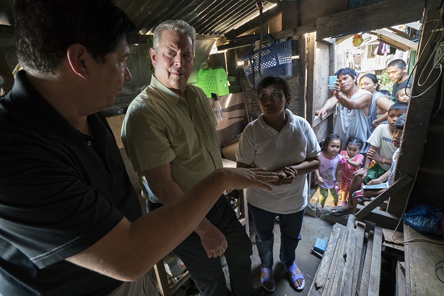 Al Gore stands in a small wooden building and speaks to two people