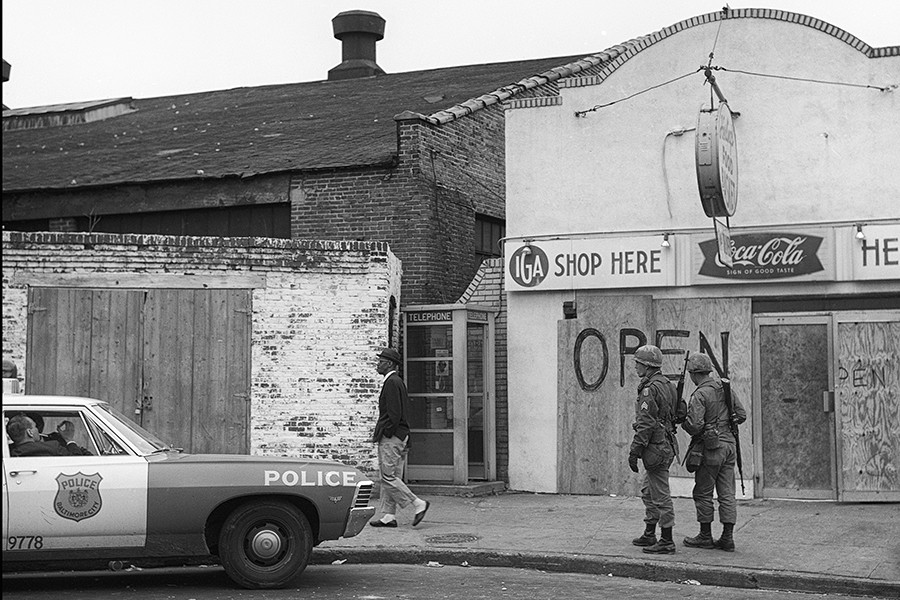 In a black and white photograph, a black man in a suit walks past boarded up shop windows, two armed soldiers, and an idle police cruiser
