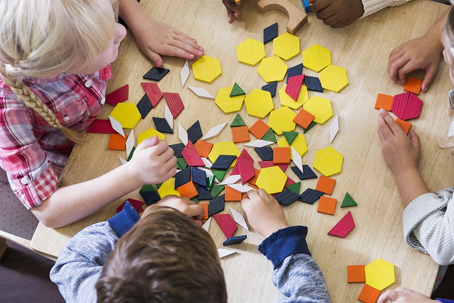 Four young kids sit at a table and play with colorful blocks in various shapes including square, hexagon, rhombus, and trapezoid