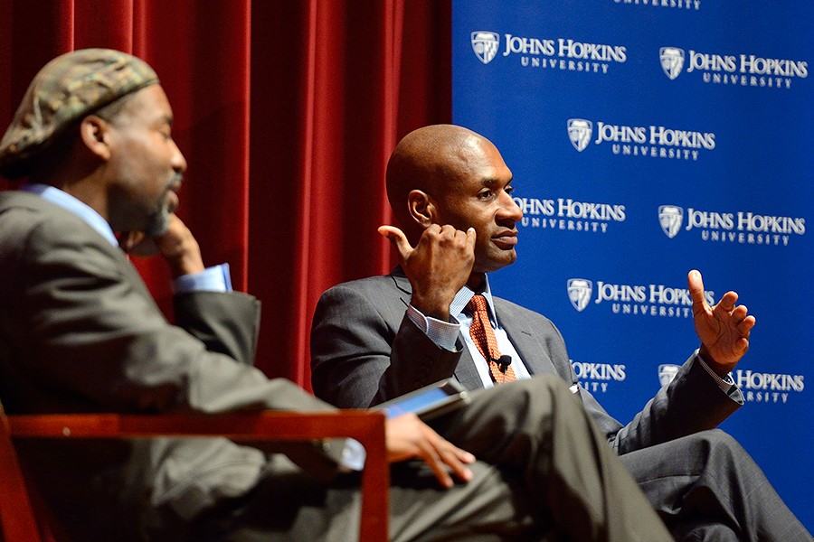 Charles Blow (right) responds to a question while seated on stage in front of a blue Johns Hopkins banner; moderator Lester Spence is seated to Blow's right