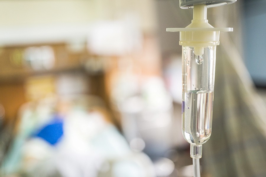 A saline drip is in the foreground while a patient in a bed is out of focus in the background