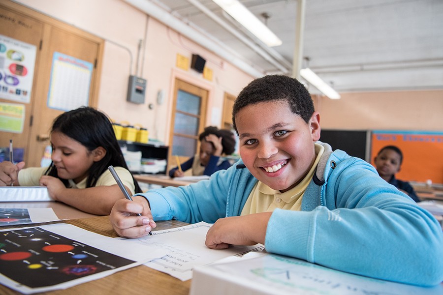 A boy smiles and looks into the camera while completing a school assignment