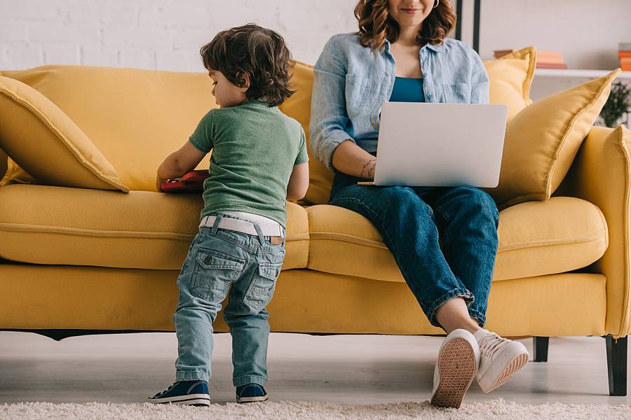 Child standing next to mother sitting on couch working on a laptop