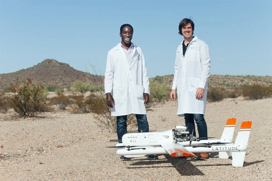 Two men in white lab coats pose next to drone in a desert setting