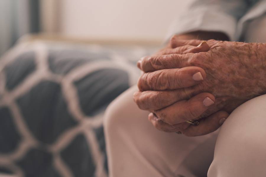An elderly person sits with their hands clasped together