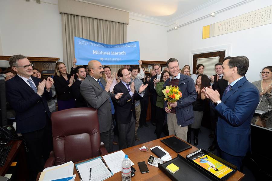A crowd of people applaud a man standing in the center of the photo holding flowers