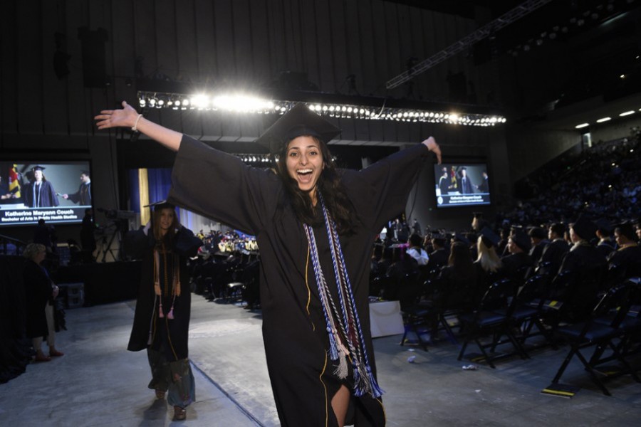 A woman in a graduation cap and gown throws her hands up triumphantly while students cross the stage behind her