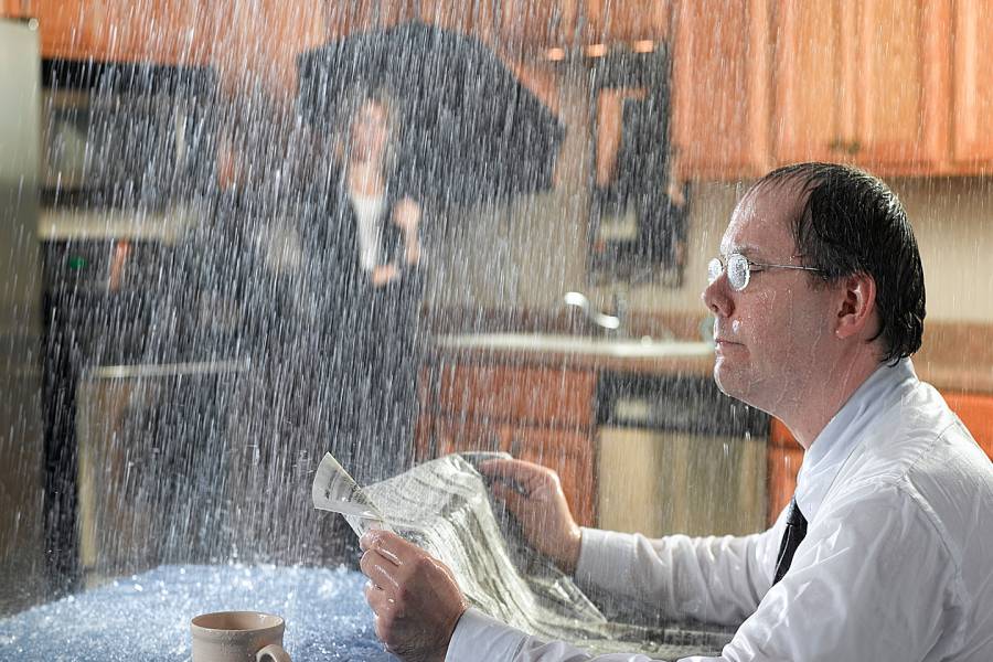 Man reading newspaper at kitchen table with water pouring through the ceiling. Woman in background is holding an umbrella.